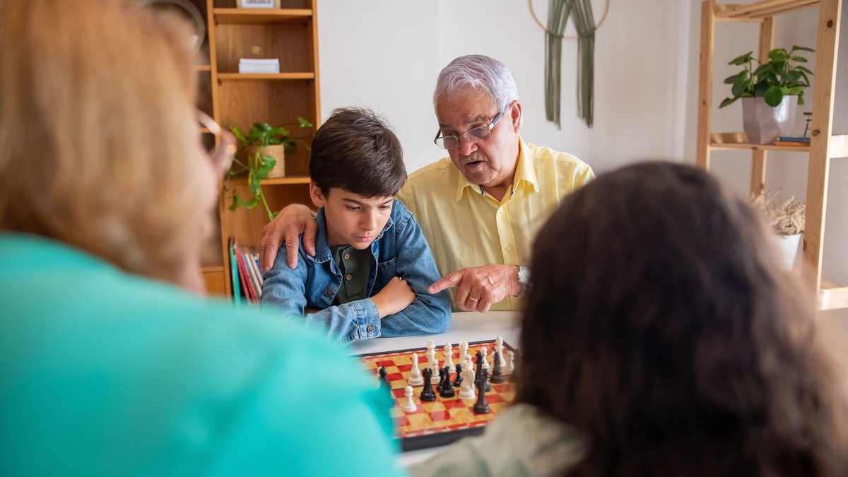 A senior man and a young boy focus intently while playing chess together indoors, surrounded by family.