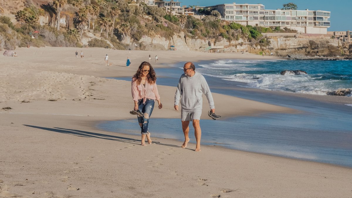 A senior couple walking barefoot along a picturesque sandy beach, enjoying a sunny day.