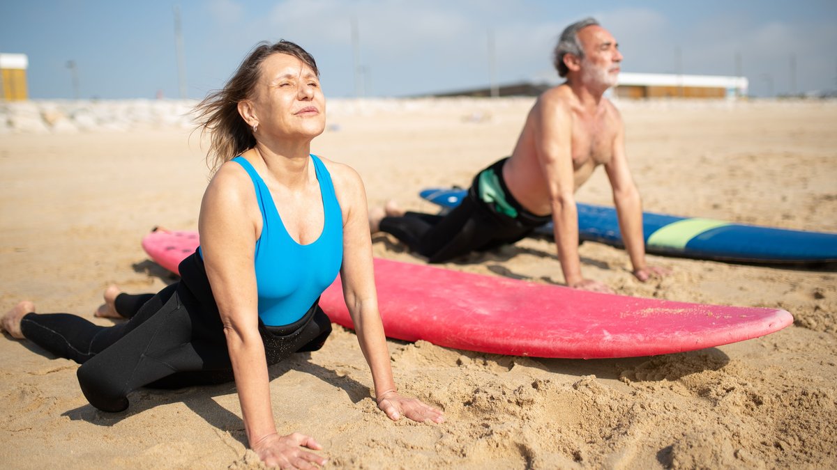 Senior couple enjoying yoga by the surfboards on a sunny beach in Portugal.
