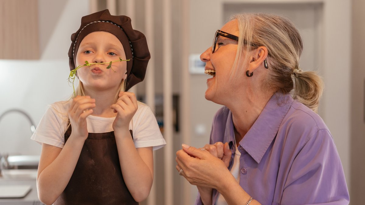 A senior woman and her young grandchild enjoy playful cooking fun indoors, sharing smiles and laughter.