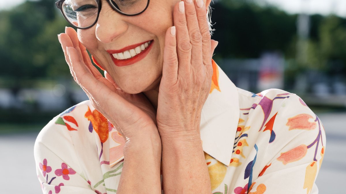 Vibrant portrait of a senior woman with glasses and a floral shirt, smiling joyfully outdoors.