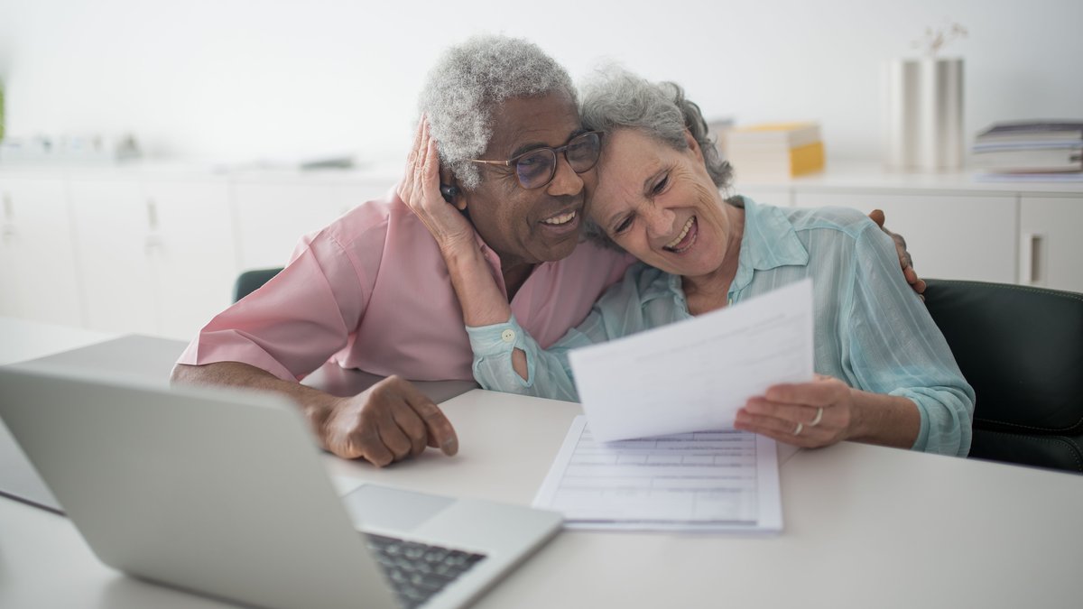 A joyful senior couple smiling while reviewing documents together at a desk with a laptop.