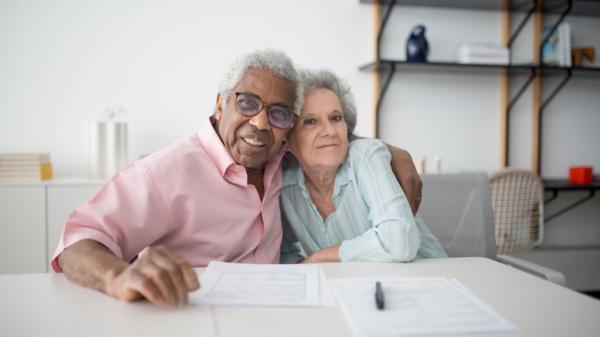 Elderly couple smiling and hugging while looking at documents on table indoors.