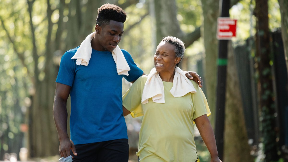 An elderly woman and young man walk arm in arm, smiling in a park. Active lifestyle.