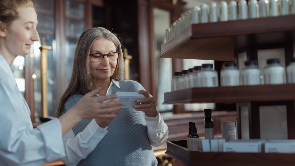 Elderly woman receiving assistance from a pharmacist in a pharmacy with product shelves.