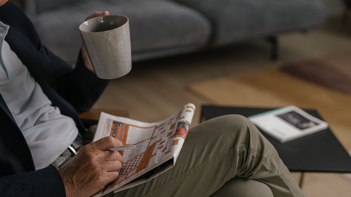 An elderly man enjoys coffee while reading a newspaper in his living room, conveying a peaceful, relaxed setting.