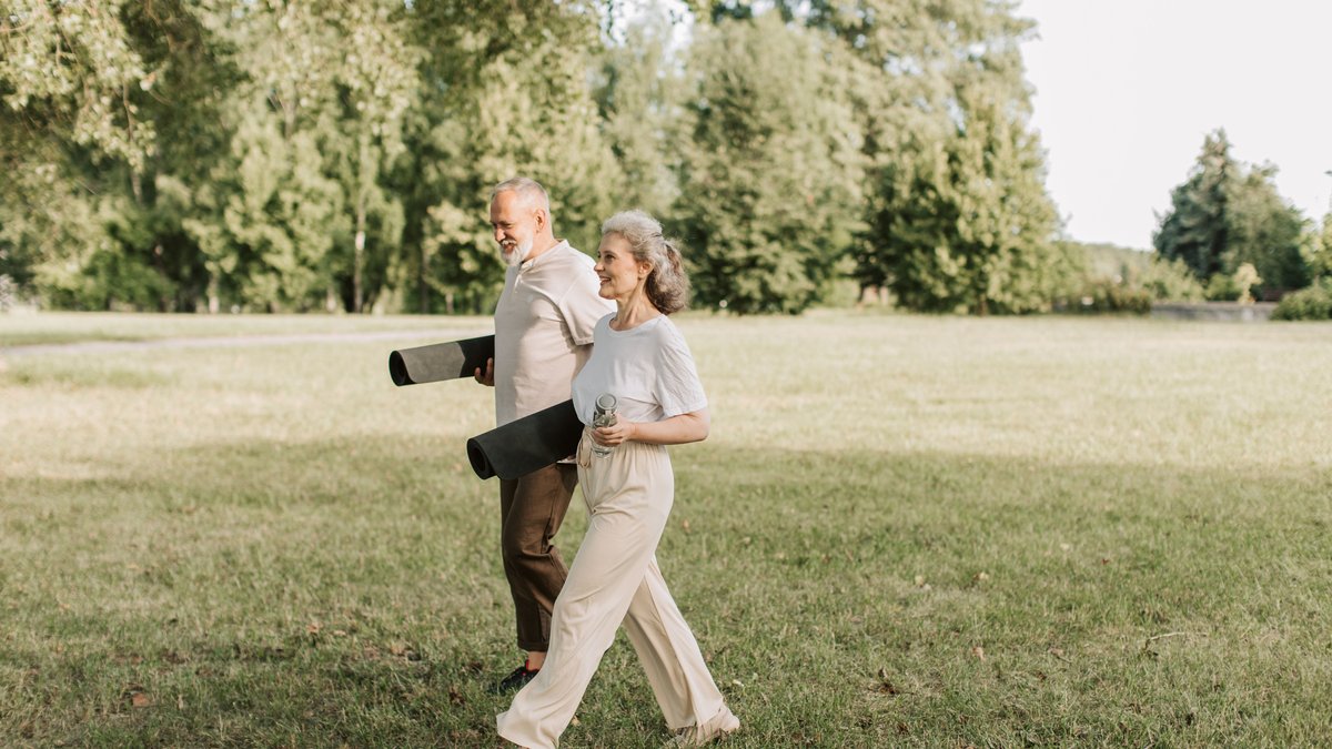 Elderly couple walking on grass carrying yoga mats, enjoying a sunny day outdoors.