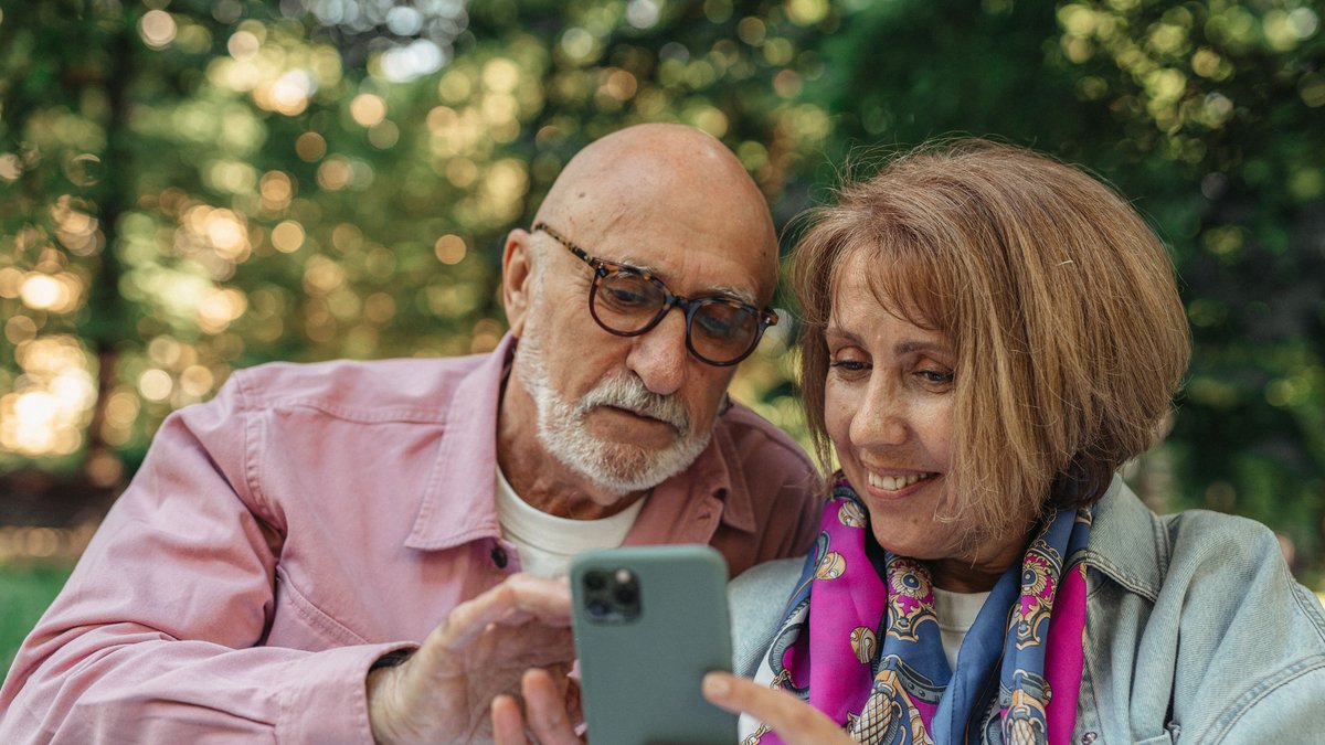 Happy senior couple using smartphone in a park, enjoying sunny outdoors.