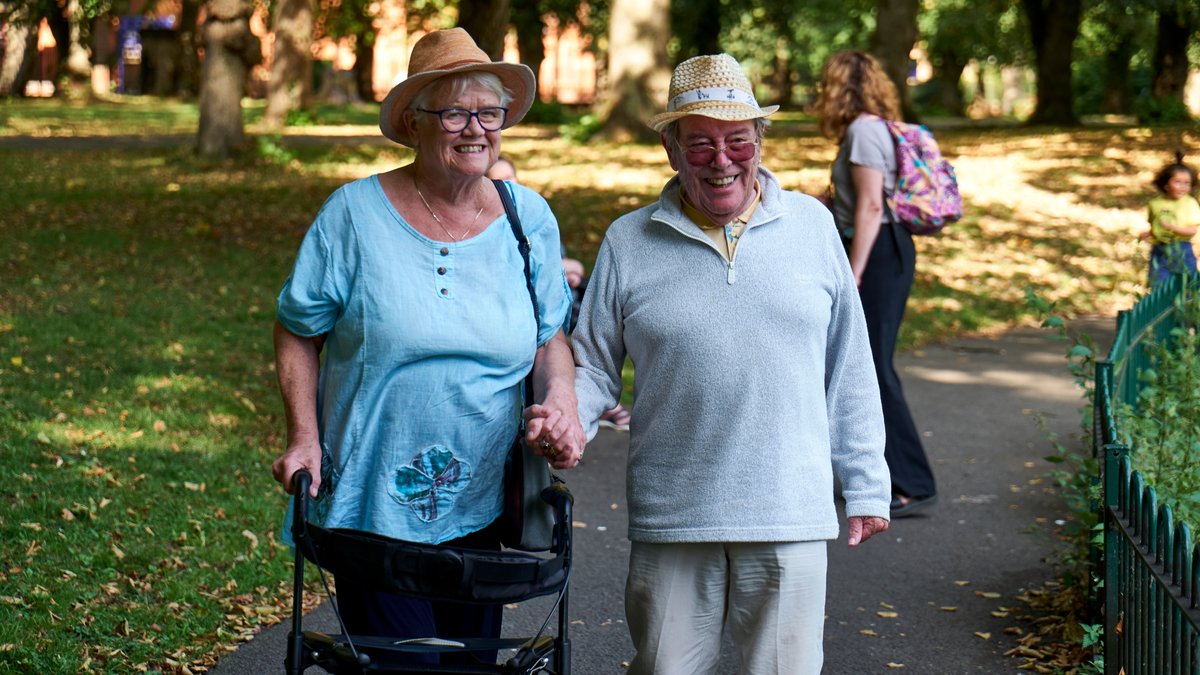 Cheerful elderly couple in casual clothes and straw hats walking on lawn of park in sunny day
